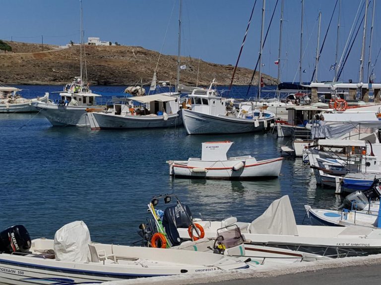 Fischerboote und Segelboote liegen dicht gedrängt im Hafen einer griechischen Kykladeninsel vor kahler Hügelküste.