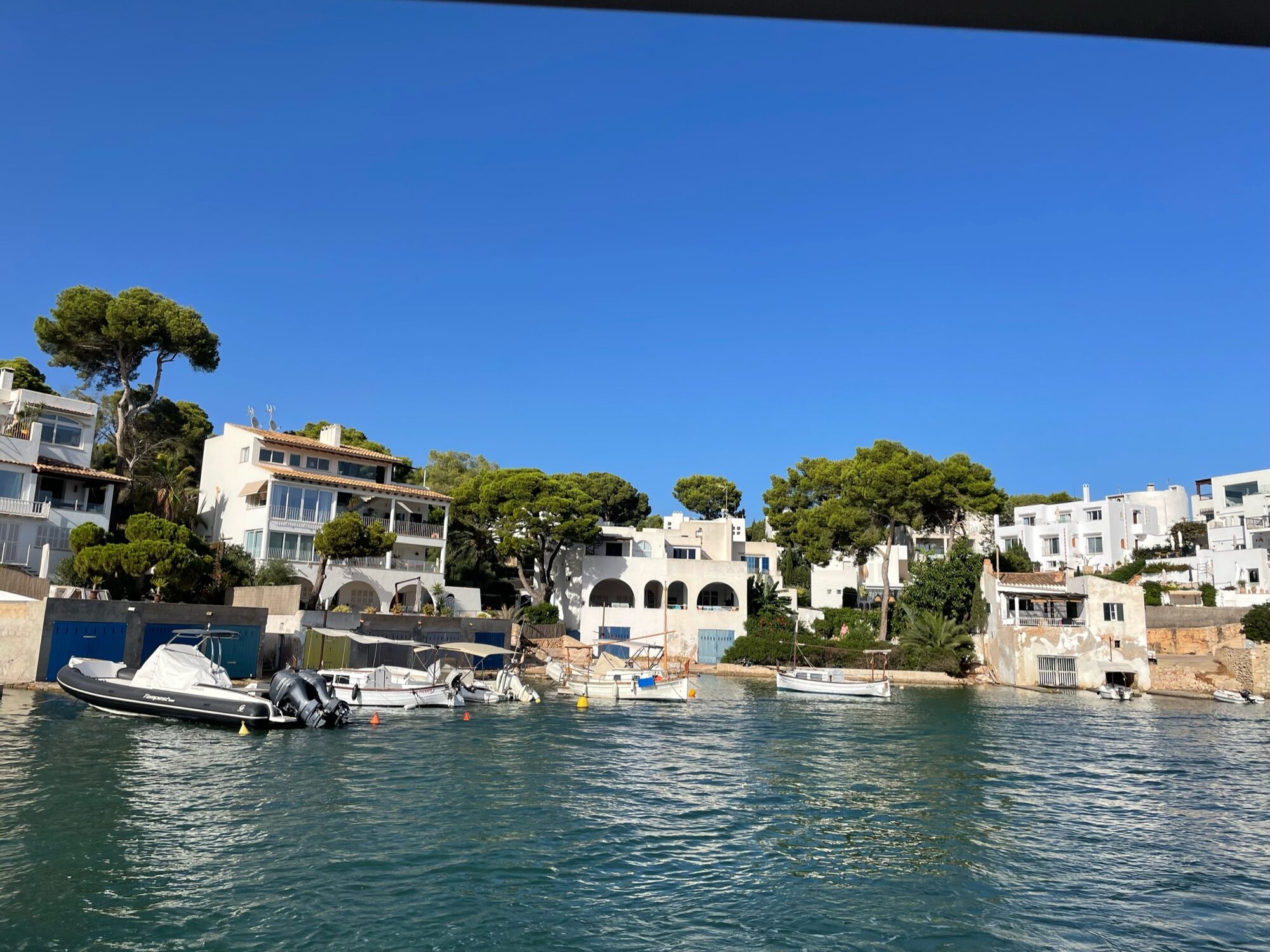Boote liegen in einer kleinen Bucht vor weißen Häusern zwischen Pinien in einem Küstenort auf Mallorca.
