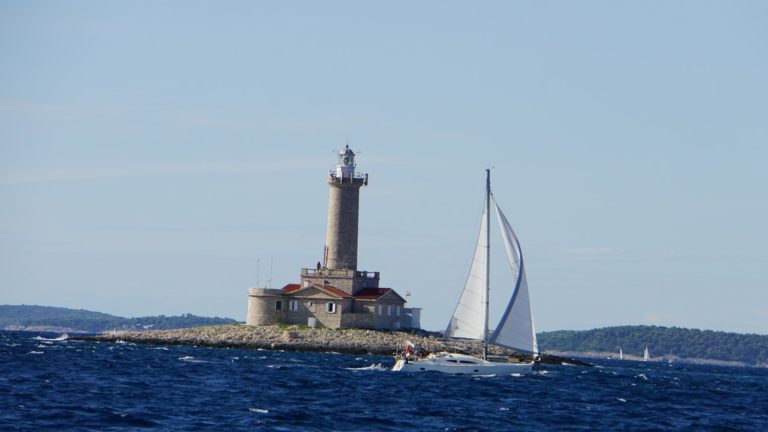 Segelboot mit gesetzten Segeln fährt an einem Leuchtturm auf felsiger Insel in der Kvarner Bucht in Kroatien vorbei.