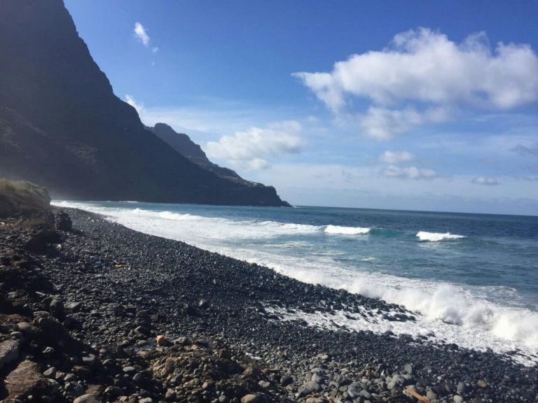 Steile Felsküste Teneriffas mit Kieselstrand, an dem Wellen des Atlantiks unter blauem Himmel mit Wolken brechen.