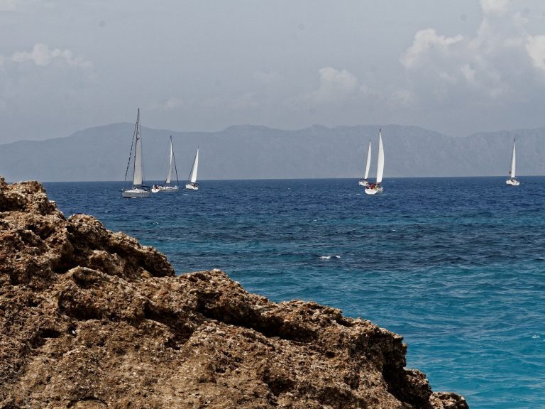 Segelboote ziehen vor einer Felsküste über das Meer vor Rhodos, im Hintergrund Berge der Ägäis.