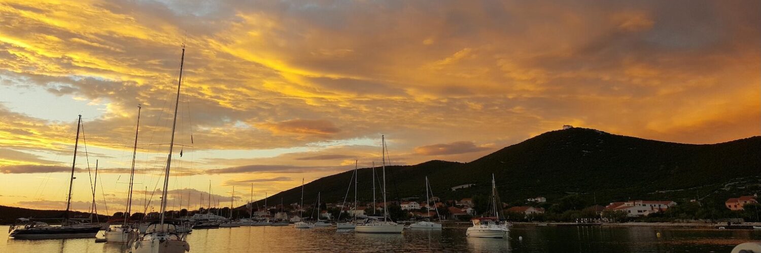 Segelboote liegen bei Sonnenuntergang in einer Bucht in Dalmatien vor einer hügeligen Küstensiedlung.