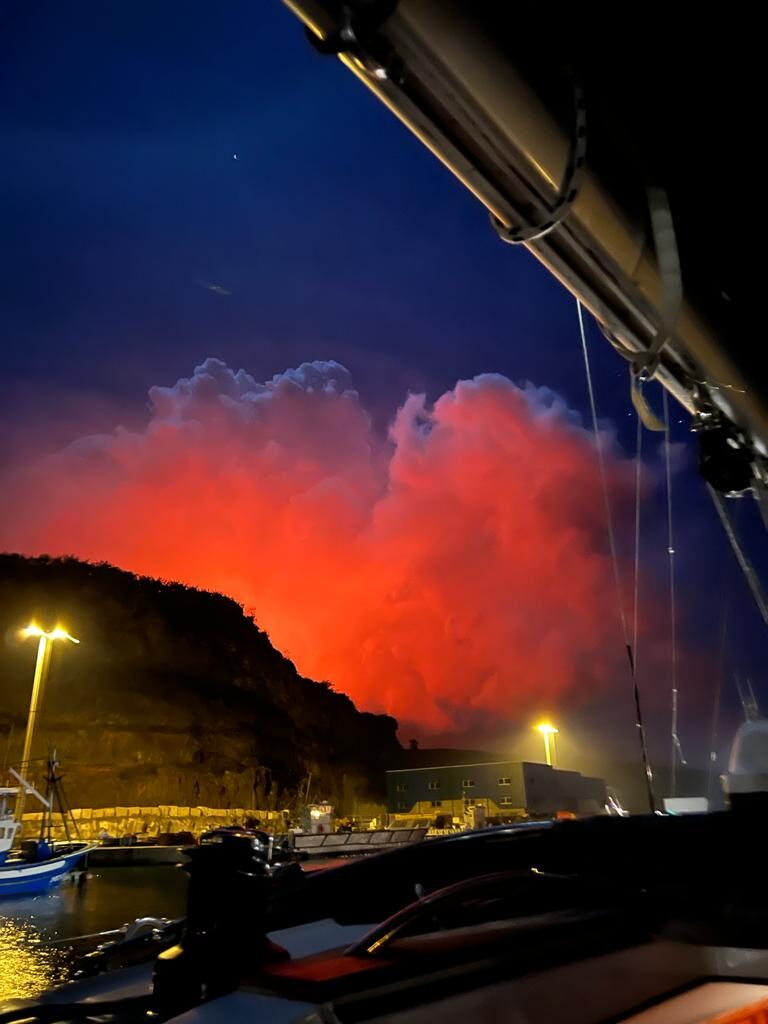 Rot erleuchtete Rauchwolke über einem Hügel, gesehen von einem Boot im Hafen von Lanzarote bei Nacht.