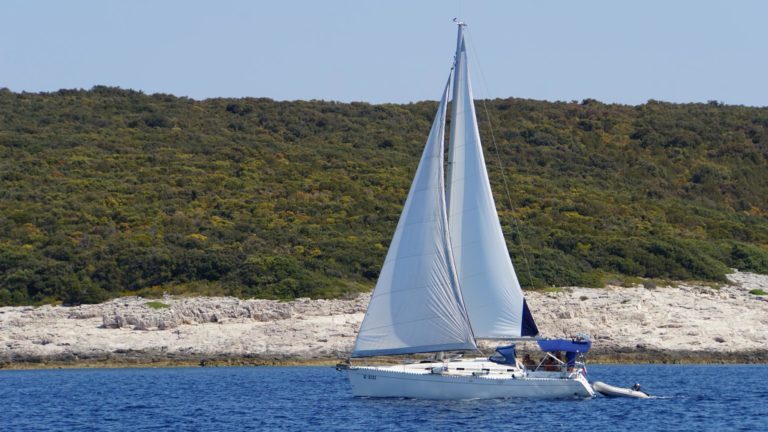 Segelboot mit gesetzten Segeln fährt an einer bewaldeten Felsküste in Kroatien vorbei.