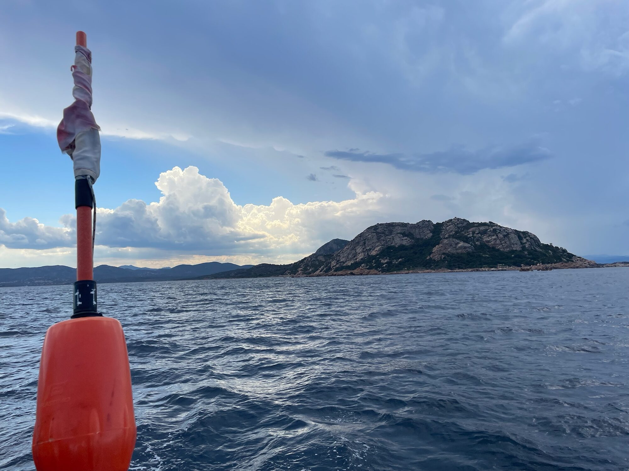 Orangefarbene Rettungsboje an Bord, im Hintergrund eine felsige Küste Sardiniens unter Wolkenhimmel.