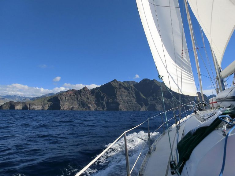 Segelboot mit gesetzten Segeln fährt vor der schroffen Felsküste Teneriffas über blaues Meer.