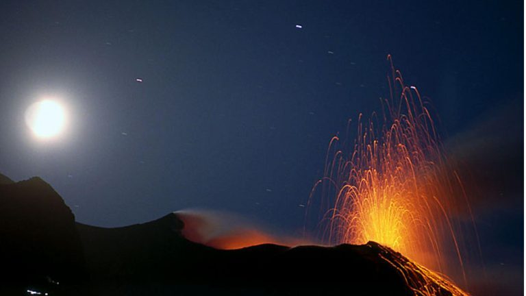 Der Vulkan Stromboli auf den Liparischen Inseln schleudert nachts glühende Lava in den Sternenhimmel neben dem Vollmond.