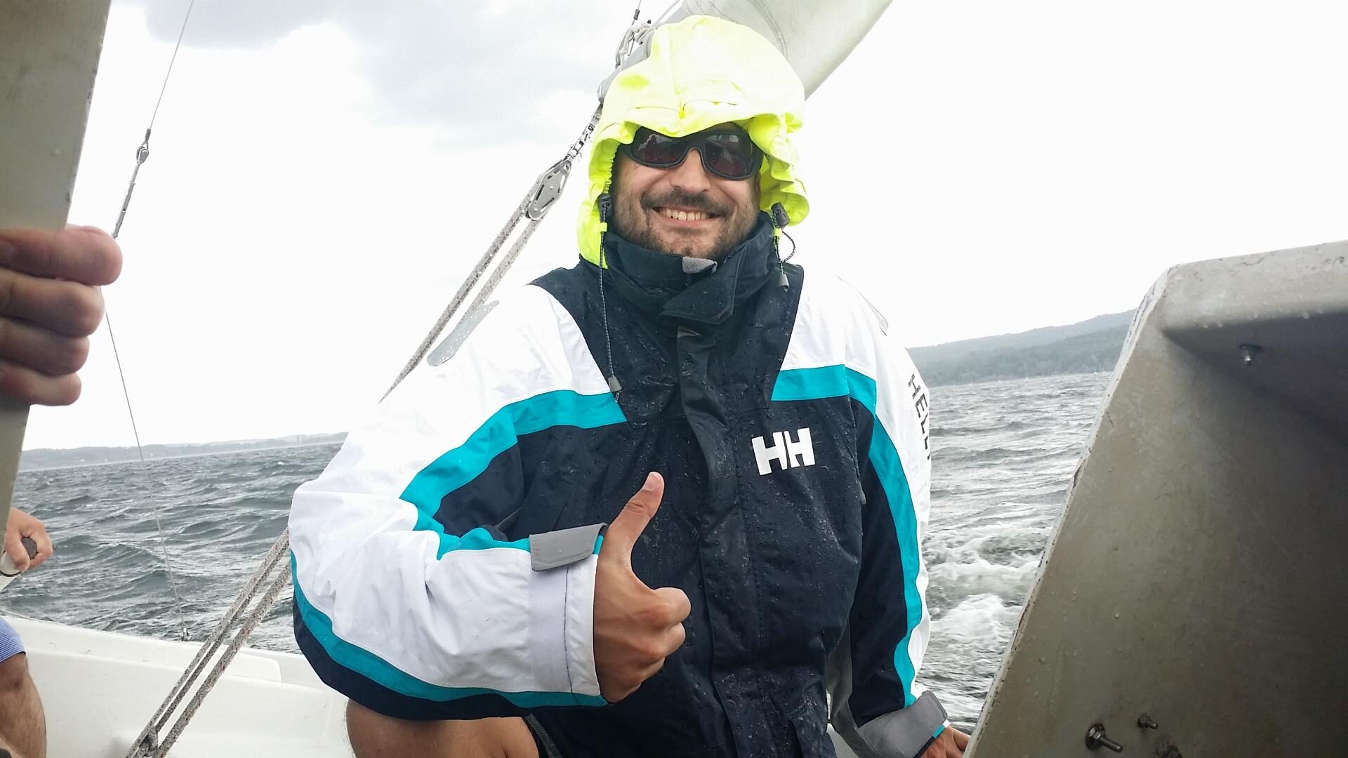 Mann in Regenjacke und gelber Kapuze zeigt Daumen hoch auf einem Segelboot im Mittelmeer.