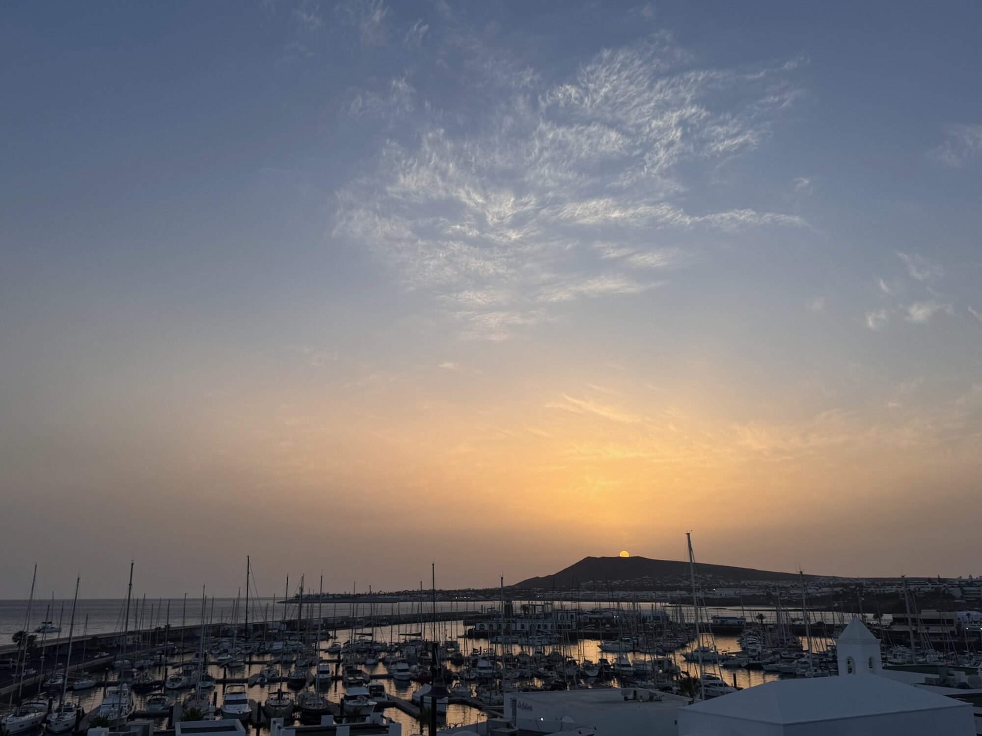 Sonnenaufgang hinter einem Vulkanhügel über der Marina Rubicón auf Lanzarote mit zahlreichen Segelbooten im Hafenbecken.
