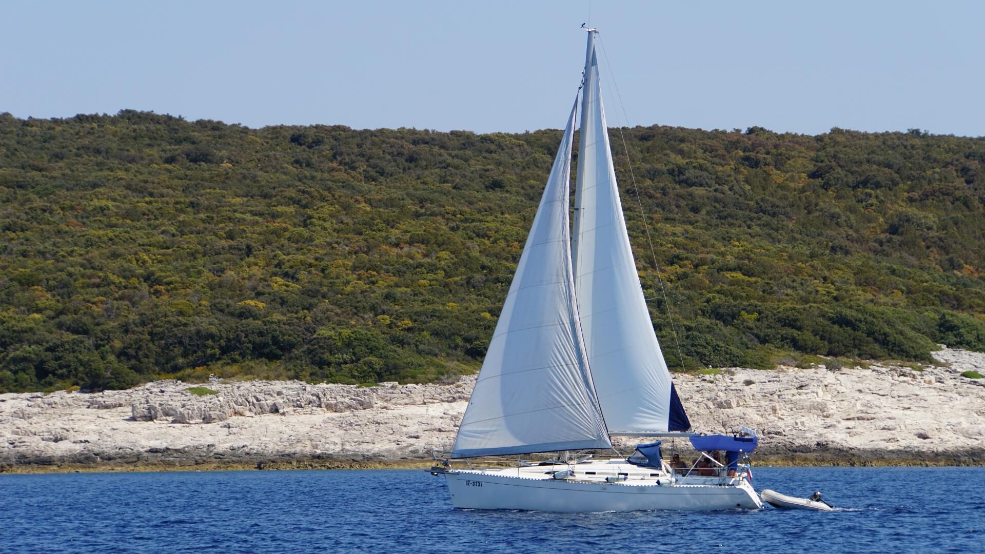 Segelboot mit gesetzter Fock und Großsegel fährt an einer bewaldeten Felsküste vorbei, ein Beiboot im Schlepp.