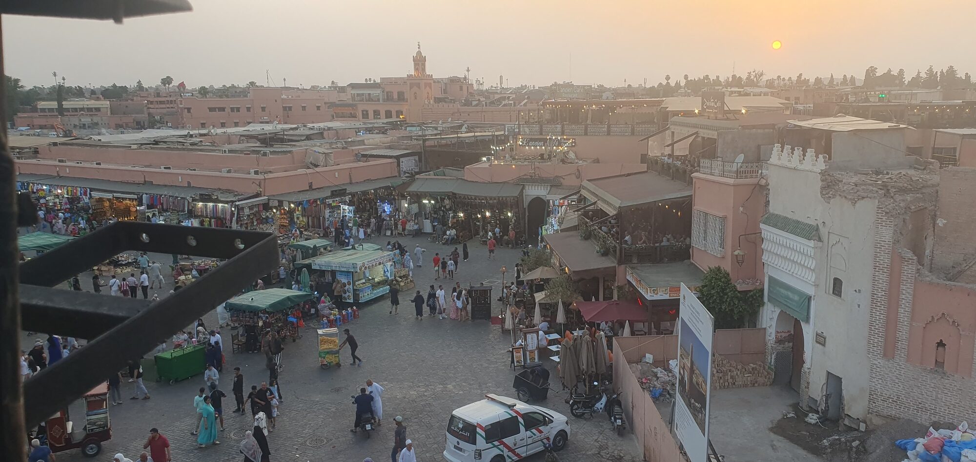 Belebter Marktplatz in Marrakesch bei Sonnenuntergang, mit Ständen, Menschen, Rikschas und einem Moscheeturm im Hintergrund.