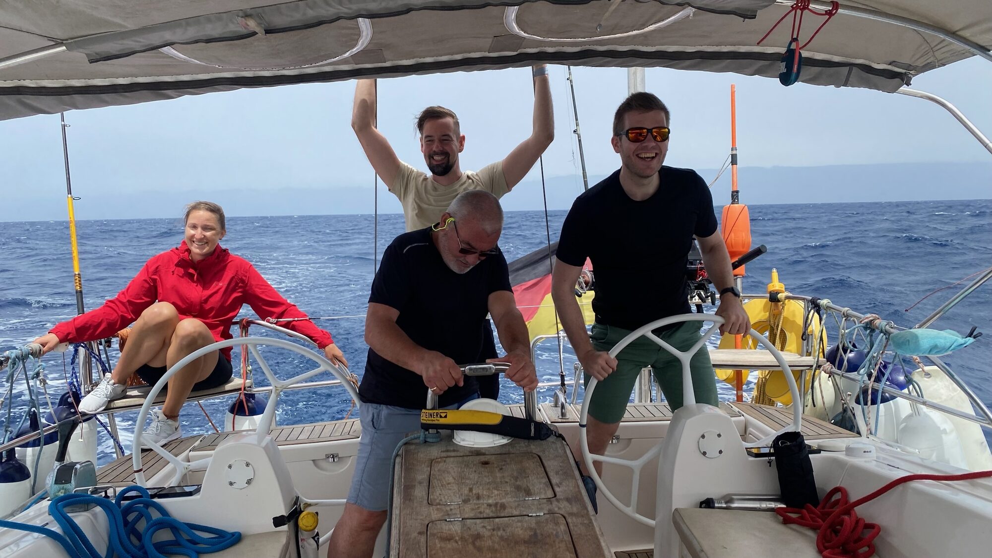 Vier lachende Segler stehen und sitzen im Cockpit einer Segelyacht auf offener See.