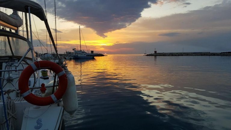 Segelboote liegen bei Sonnenuntergang in einem Hafenbecken vor Sardinien, im Vordergrund ein Rettungsring am Bug.
