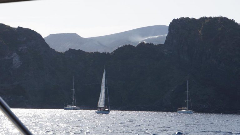 Segelboote gleiten vor schroffen Felsküsten der Liparischen Inseln über das glitzernde Meer.