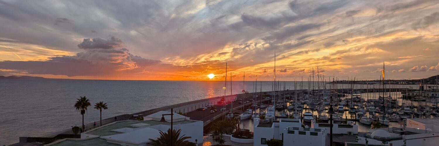 Sonnenuntergang über der Marina Rubicón auf Lanzarote, mit Segelbooten am Steg und Palmen am Ufer.