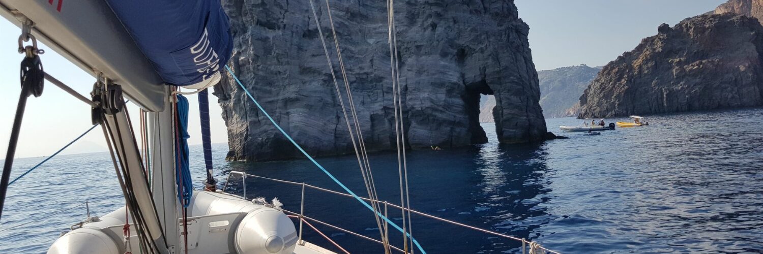 Segelboot vor einer Felsformation mit natürlichem Bogen im Meer bei den Liparischen Inseln, im Hintergrund kleine Boote.