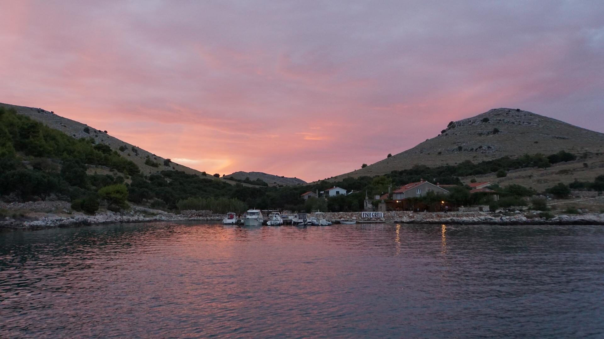 Bucht der Kornaten bei Sonnenuntergang mit Booten am Steg vor einem Fischrestaurant zwischen kahlen Hügeln.