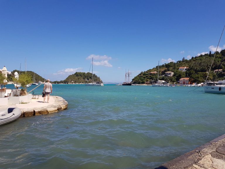 Eine Person steht an einer Kaimauer vor einer türkisblauen Bucht mit Segelbooten und grünen Hügeln im Ionischen Meer.