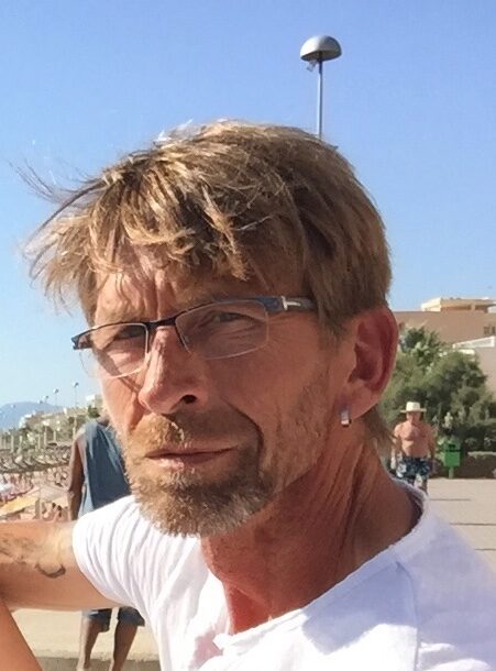 Helge Brunke mit Brille und weißem Shirt an einer Strandpromenade bei ActionTeam-Sailing.