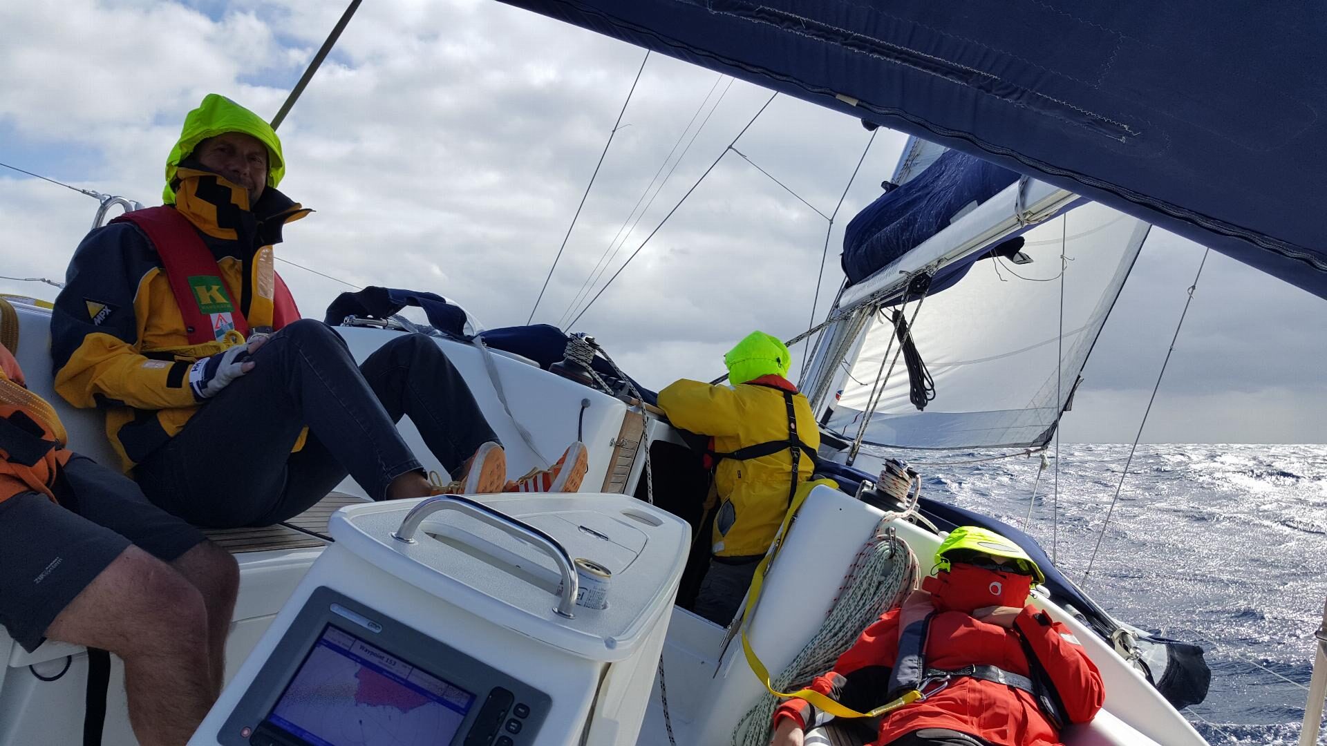Crew in gelber und roter Ölzeug-Regenkleidung sitzt an Bord eines Segelboots auf offener See.