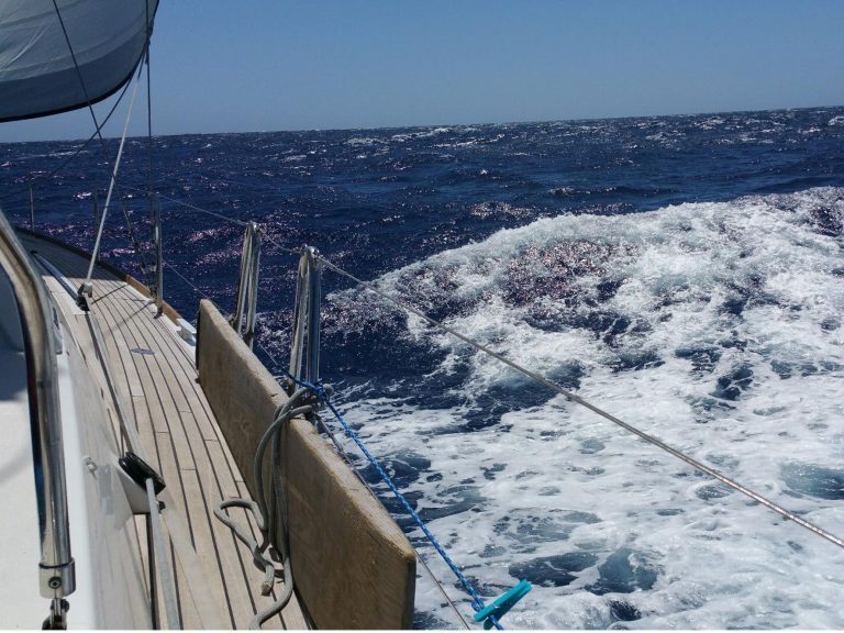 Segelboot mit Teakdeck pflügt durch tiefblaue Wellen auf dem Atlantik zwischen Kanaren und Madeira, weiße Gischt am Heck.