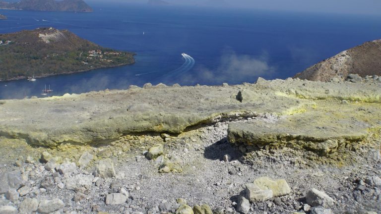 Schwefelablagerungen am Kraterrand von Vulcano, Blick auf Buchten, ein Boot und weitere Liparische Inseln im Meer.