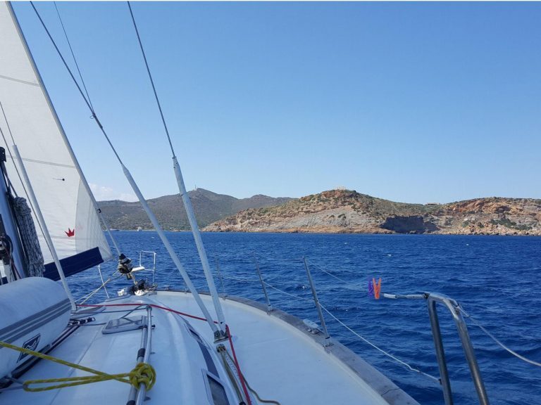 Segelboot fährt an einer felsigen Küste mit Tempelruine auf einer Anhöhe in den Kykladen vorbei.