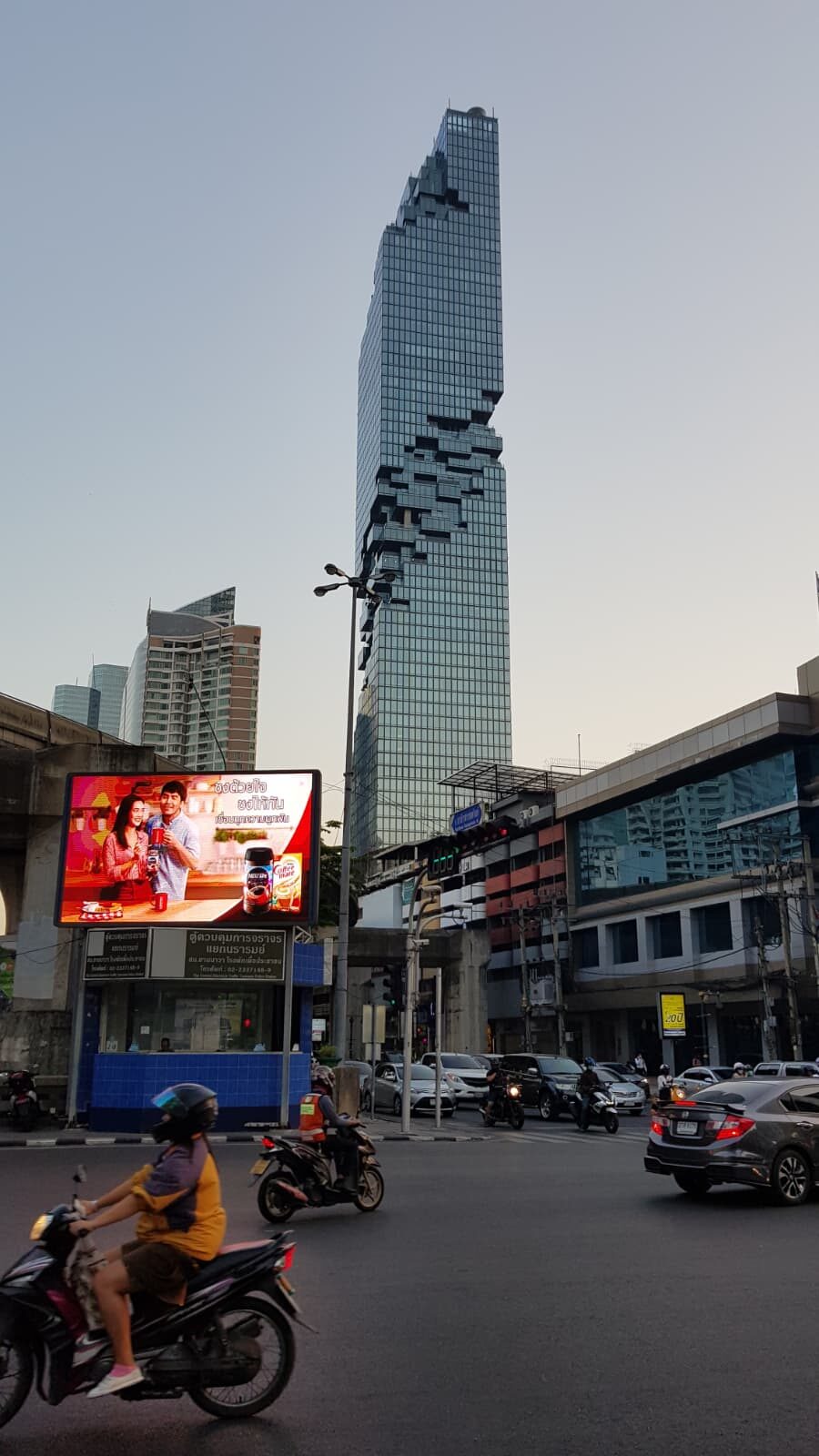Wolkenkratzer MahaNakhon in Bangkok, im Vordergrund Straßenverkehr mit Motorrollern, Autos und einer Werbetafel.