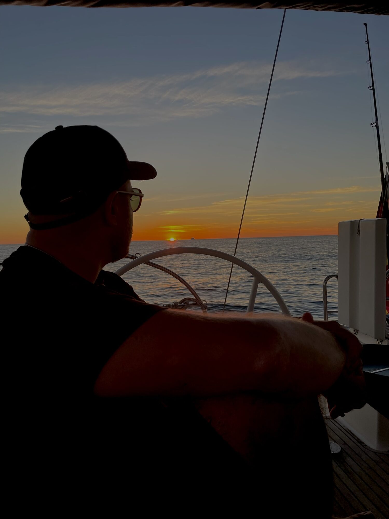 Ein Mann sitzt am Steuerrad eines Segelboots auf dem Meer und blickt bei Sonnenuntergang zum Horizont.