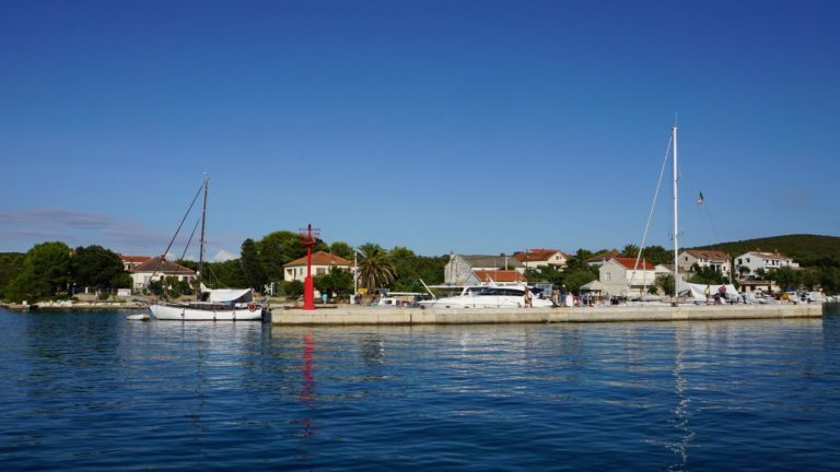 Segelboote liegen an einer Hafenmole vor einem kroatischen Küstendorf mit roter Leuchtbake.