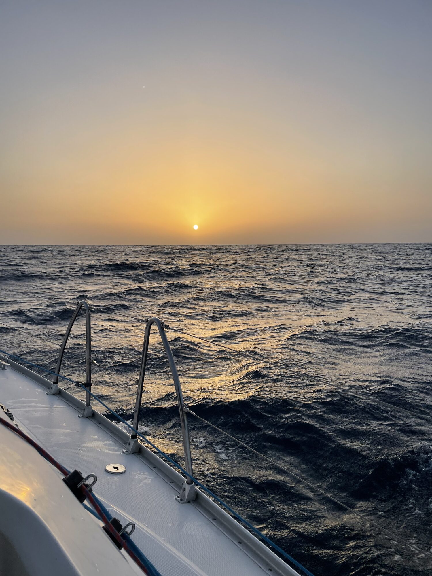Sonnenuntergang über bewegter See, vom Deck eines Segelboots mit Relingstützen aus gesehen.