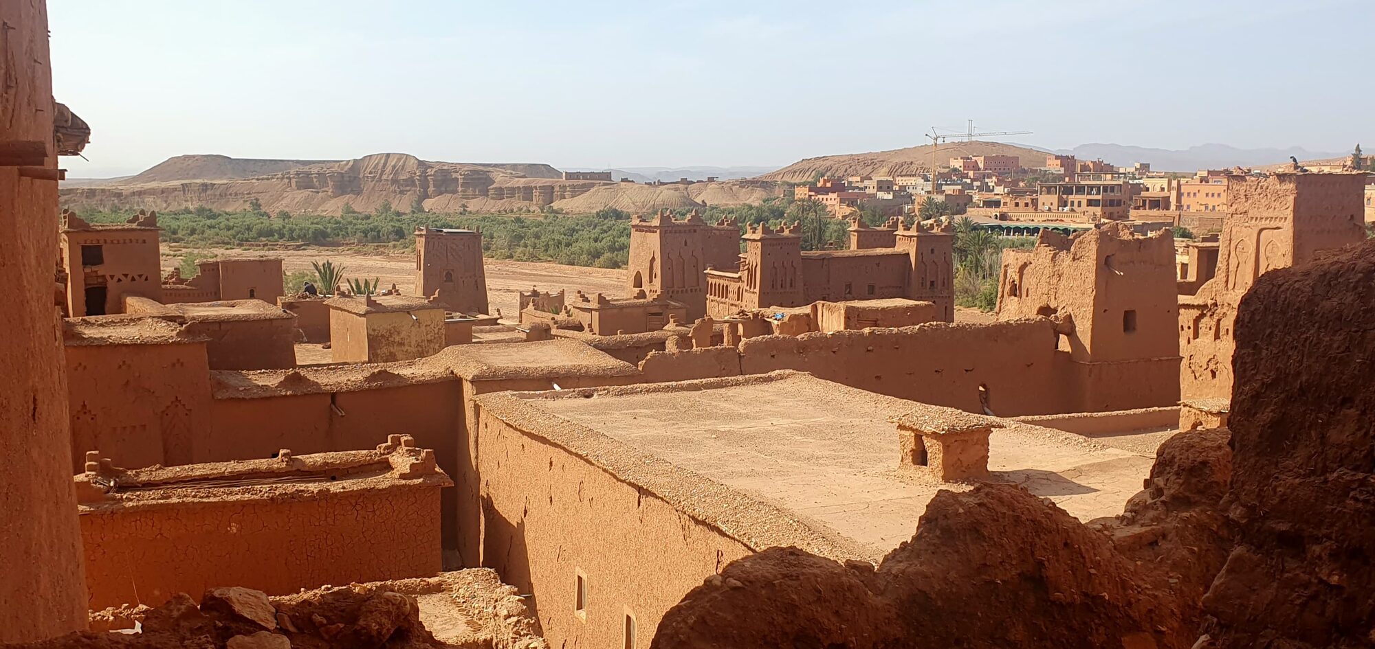 Blick über die Lehmziegelbauten von Aït Benhaddou mit Türmen, Palmenoase und Bergen im Hintergrund.
