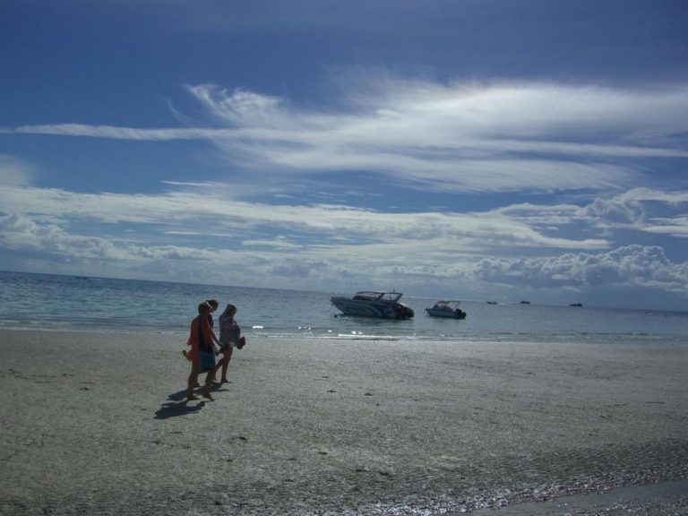 Zwei Personen gehen an einem Strand bei Krabi, im flachen Wasser dahinter liegen zwei Motorboote vor Anker.