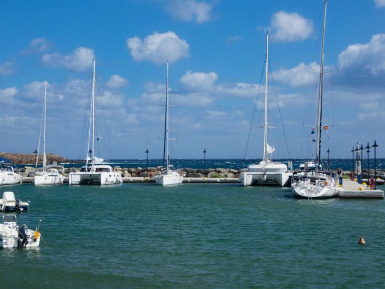 Segelboote und Katamarane liegen an einem Steg in einem griechischen Hafen unter blauem Himmel.