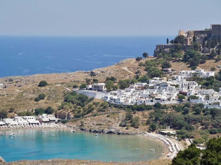 Weiße Häuser von Lindos auf Rhodos liegen zwischen Akropolis-Festung auf dem Hügel und türkiser Bucht mit Sandstrand.