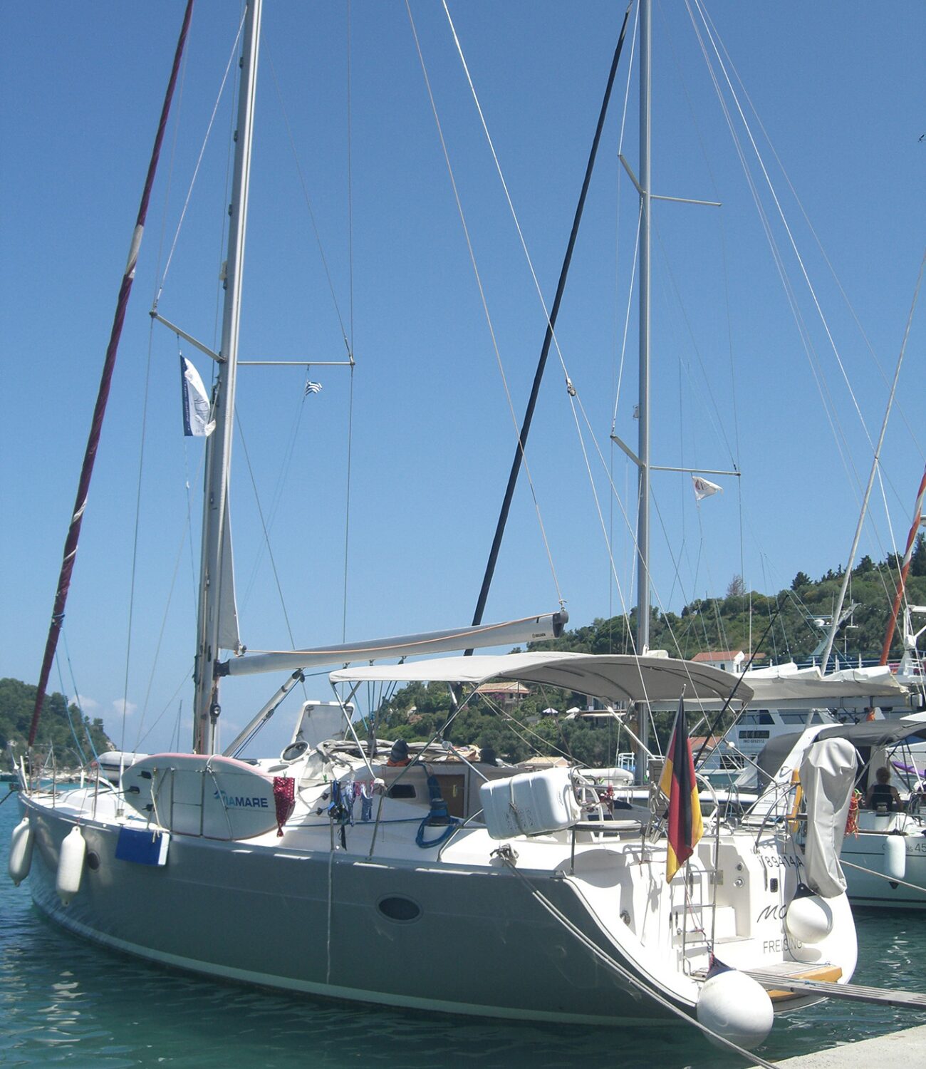 Segelboot mit deutscher Flagge liegt im Hafen an einem Steg, im Hintergrund grüne Hügel mit Häusern.