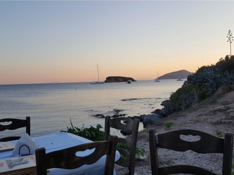 Segelboote ankern vor einer felsigen Bucht in Griechenland, gesehen von einer Taverne am Ufer bei Sonnenuntergang.