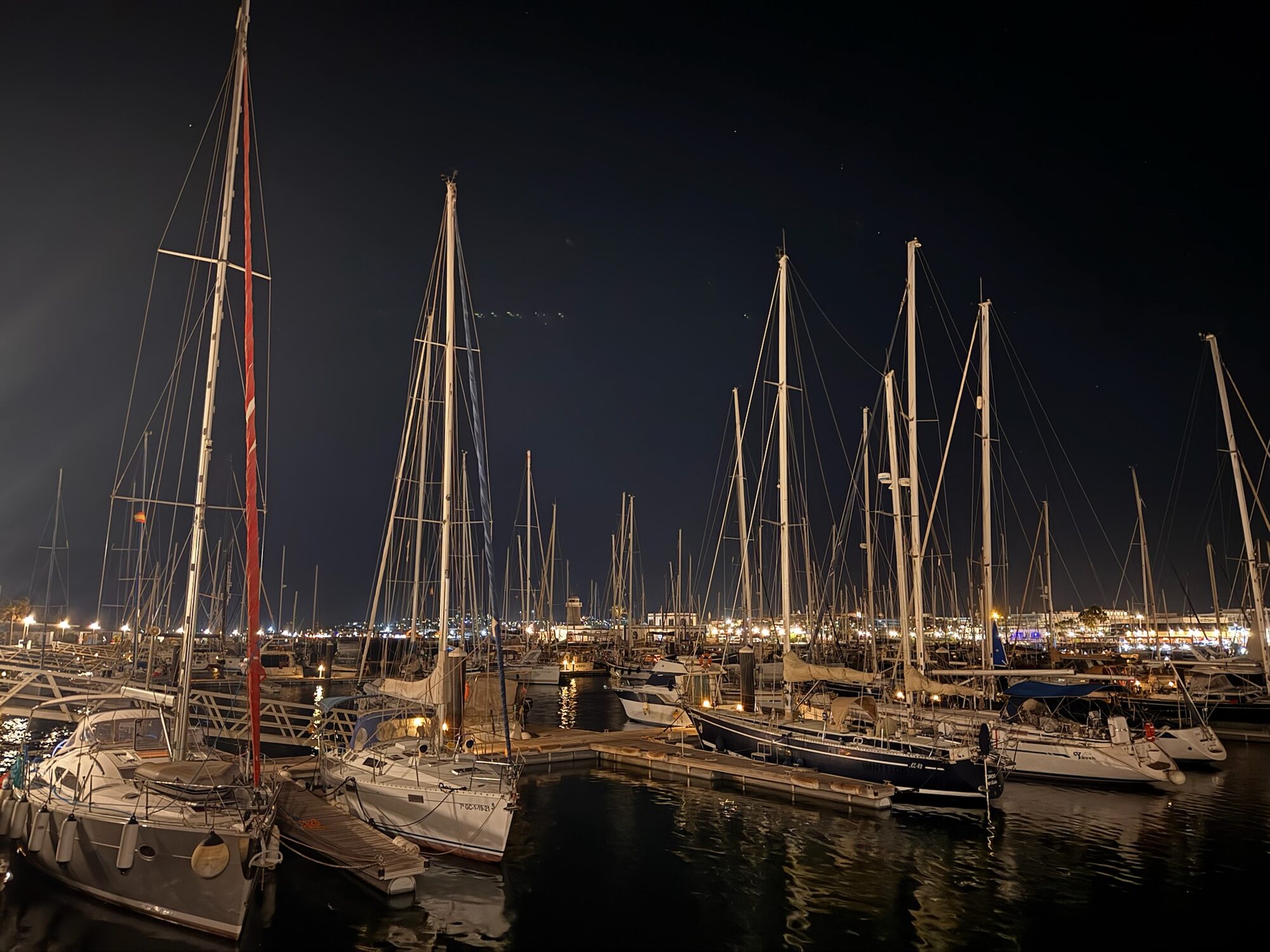 Segelboote liegen bei Nacht an Stegen in der Marina Rubicón auf Lanzarote, im Hintergrund beleuchtete Uferpromenade.