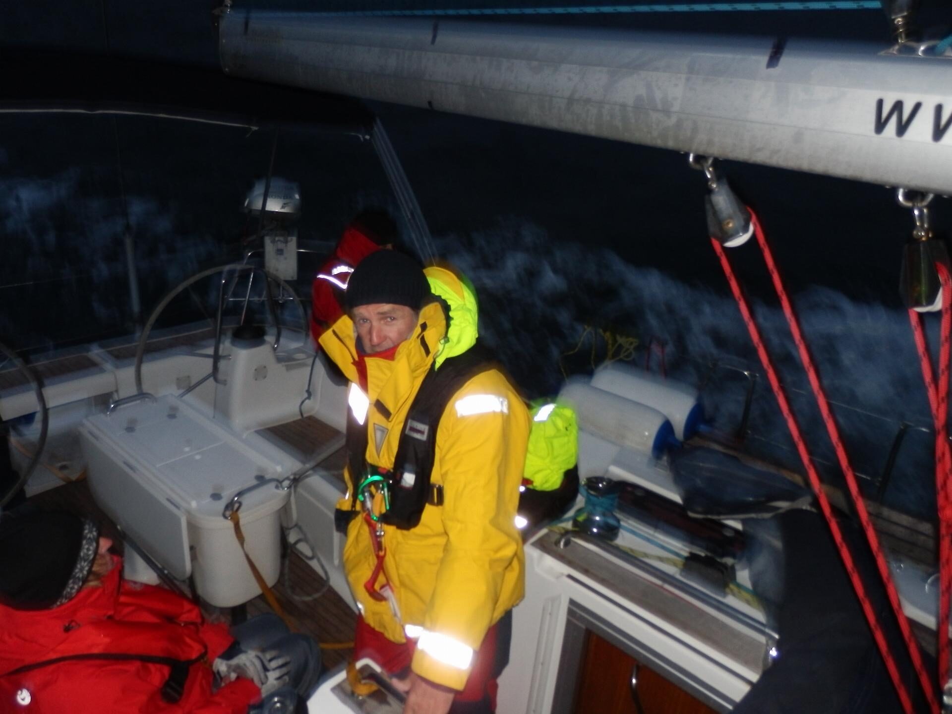 Segler in gelber Ölzeugjacke mit aufgeblasener Rettungsweste steht bei Nacht im Cockpit eines Segelboots.