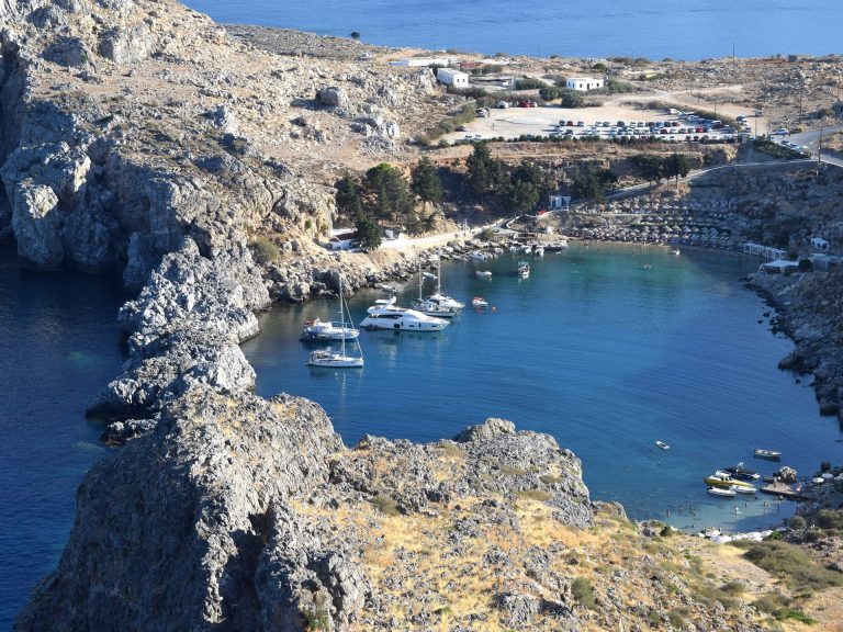 Felsige Bucht auf Rhodos mit ankernden Segel- und Motoryachten, umgeben von Klippen und einem Parkplatz am Ufer.