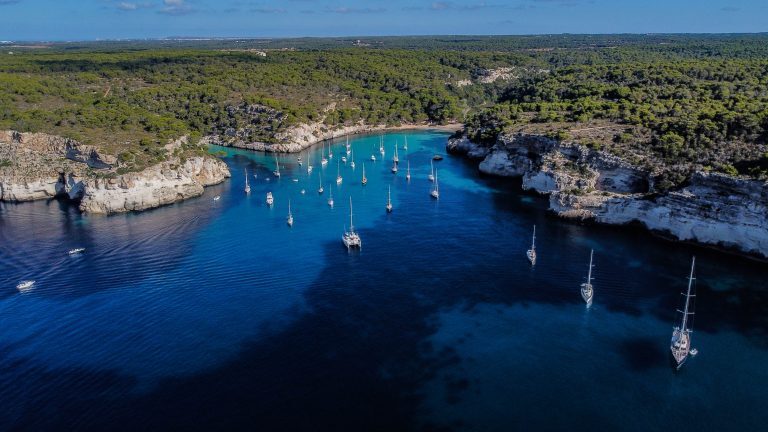 Segelboote ankern in einer türkisblauen Bucht zwischen bewaldeten Felsklippen an der Küste.