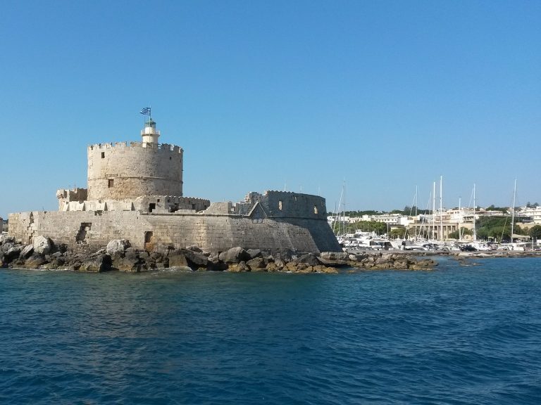 Festung Agios Nikolaos am Hafeneingang von Rhodos-Stadt, mit griechischer Flagge auf dem Turm und Segelbooten im Hintergrund.