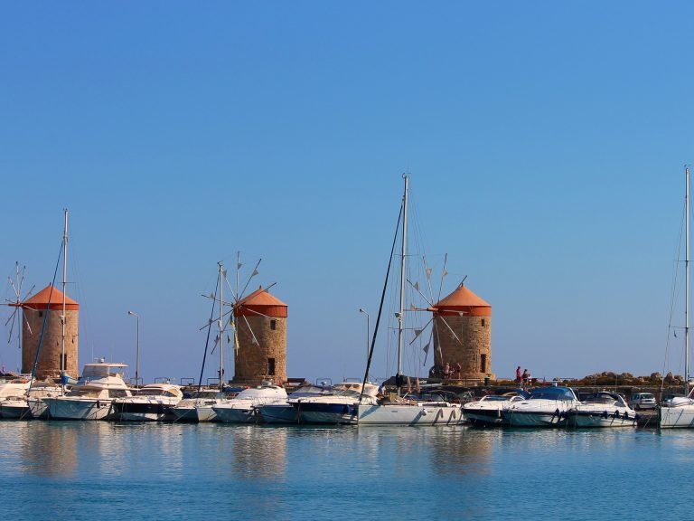 Windmühlen am Hafen von Rhodos-Stadt, davor liegen Segel- und Motorboote im ruhigen Wasser.