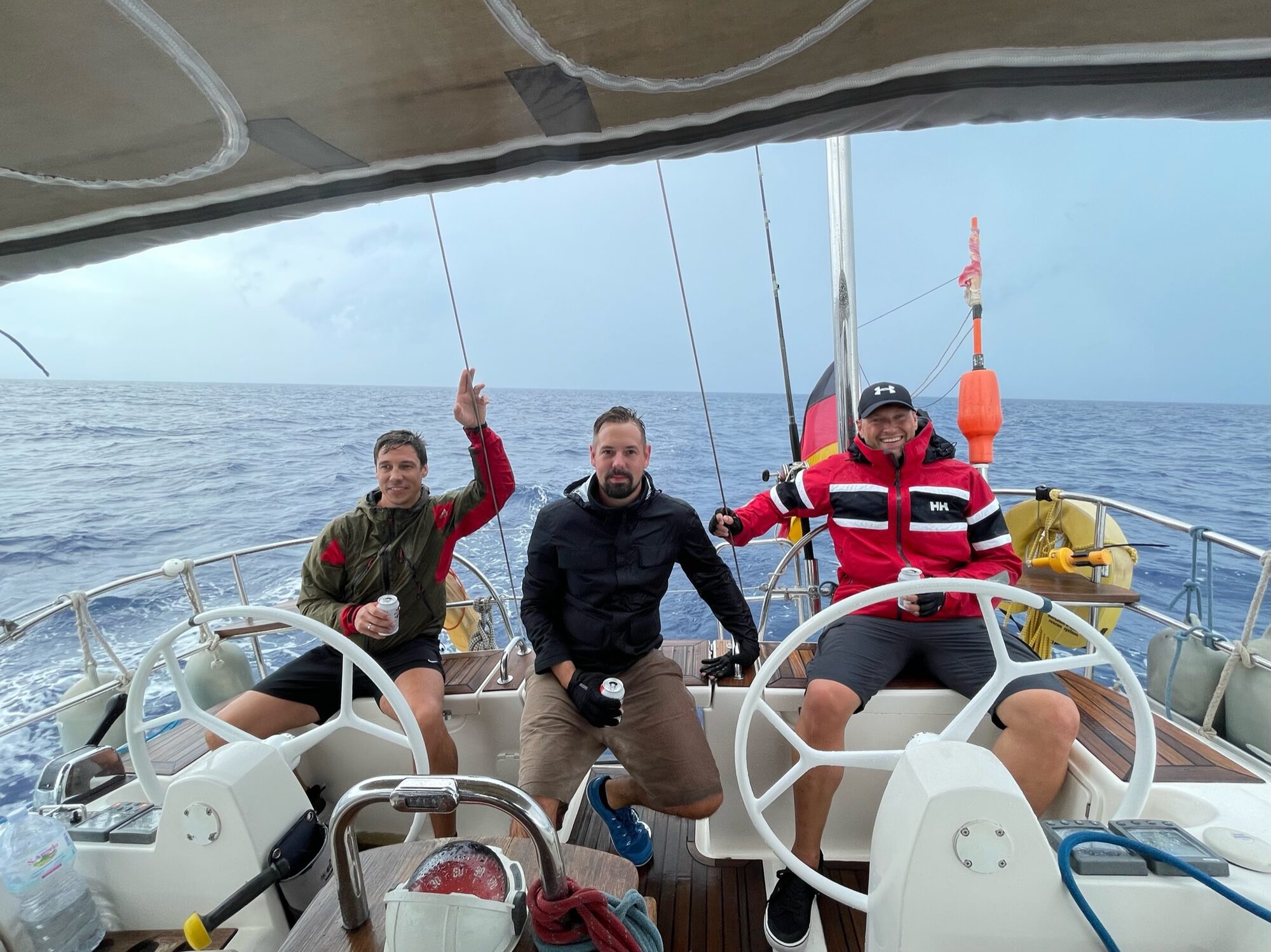 Drei Männer sitzen mit Getränken im Cockpit eines Segelboots vor den Kanarischen Inseln und winken in die Kamera.