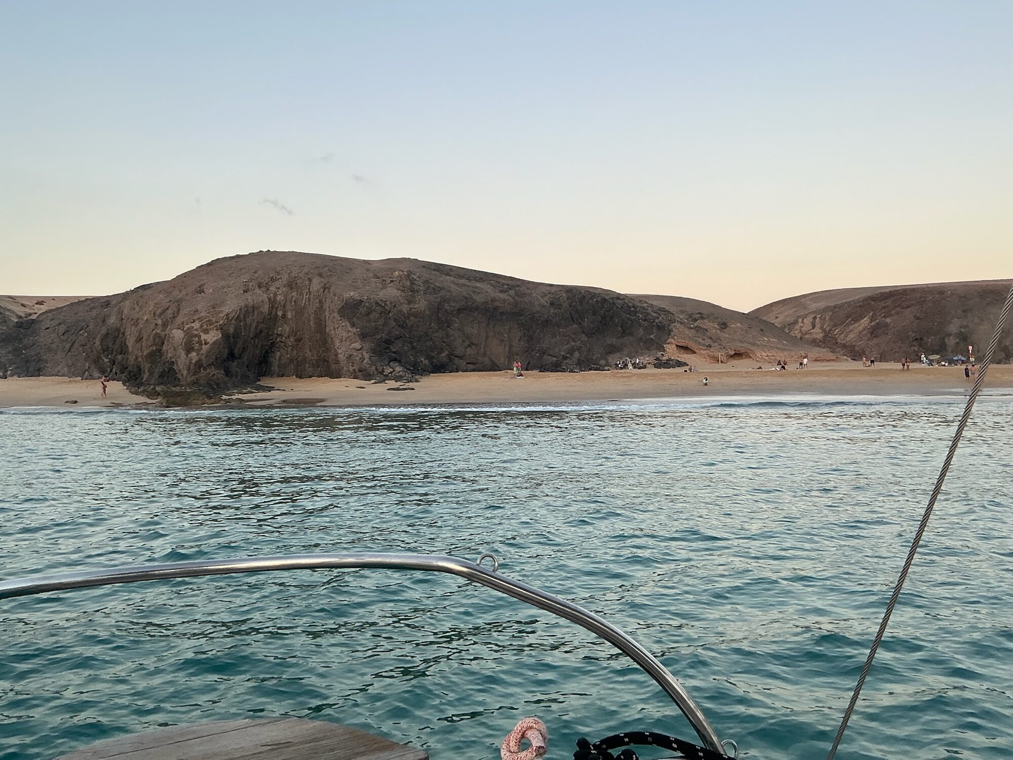 Blick vom Bootsdeck über das Wasser auf einen Sandstrand mit Felsen vor Lanzarote, Menschen am Ufer.
