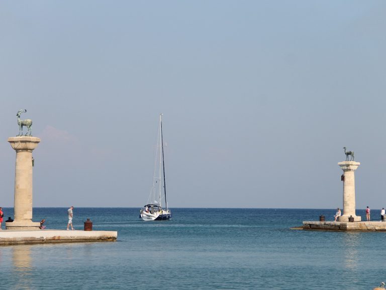 Segelboot fährt zwischen den Säulen mit Hirsch- und Hirschkuhstatuen in die Hafeneinfahrt von Rhodos-Stadt.