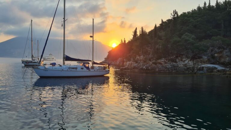 Segelboote liegen bei Sonnenuntergang vor einer bewaldeten Felsküste in einer ruhigen Bucht.