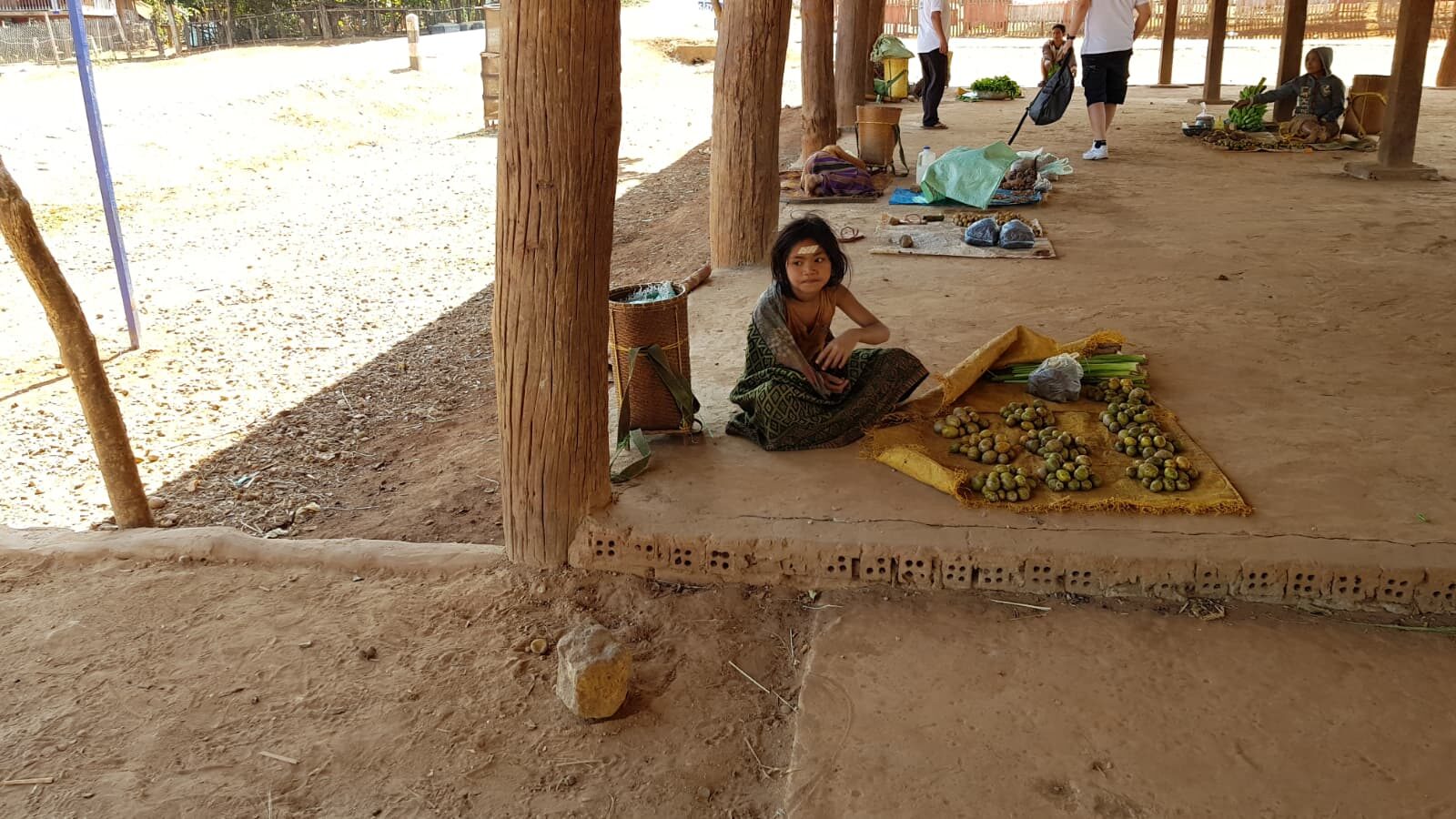 Kind sitzt auf Markttuch unter offenem Holzpavillon in Thailand, vor sich ausgebreitete grüne Früchte auf einer Matte.