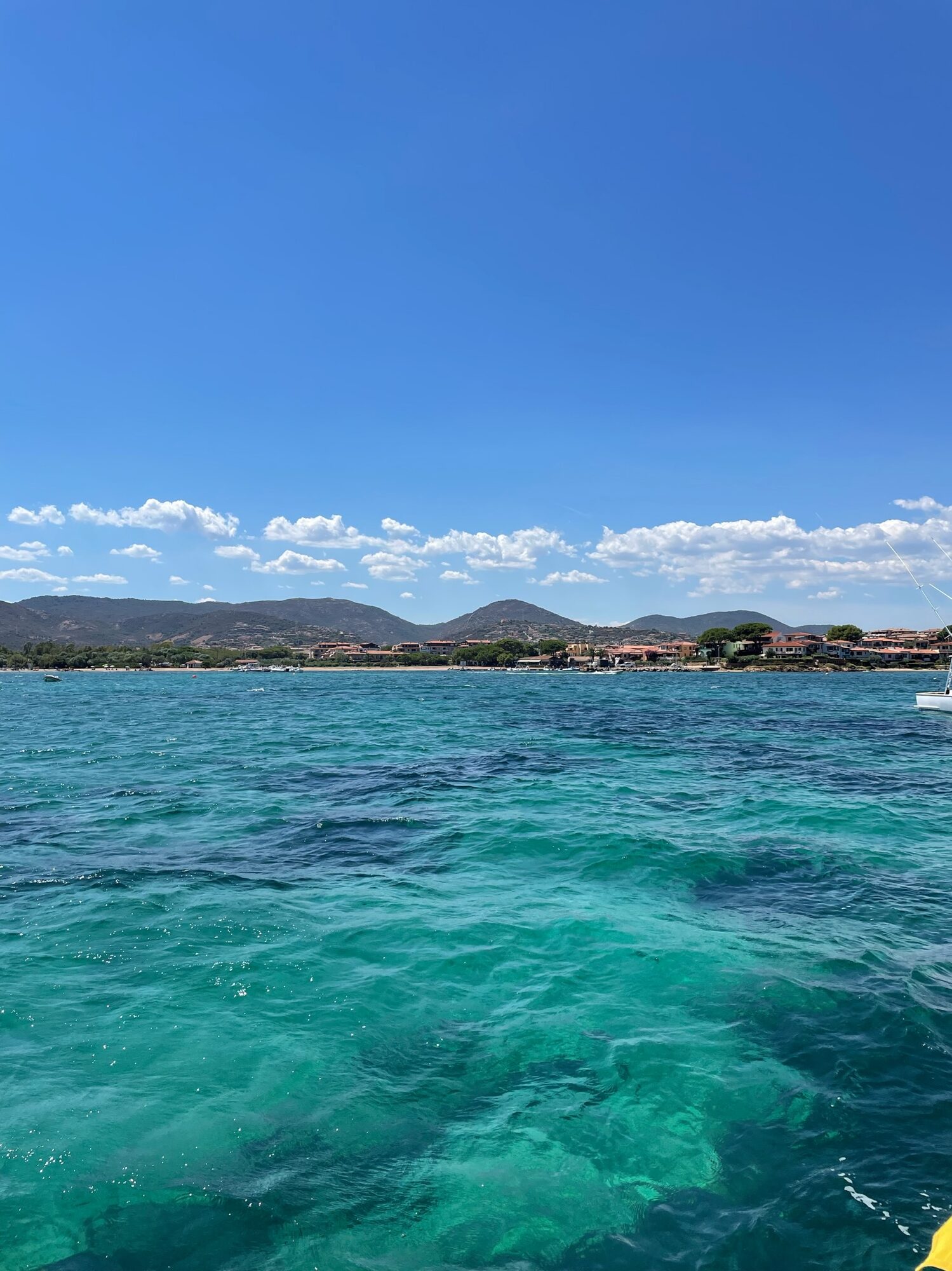 Türkisfarbenes Meer vor einer Küstenstadt mit Hügeln im Hintergrund, am rechten Bildrand ein Segelboot.