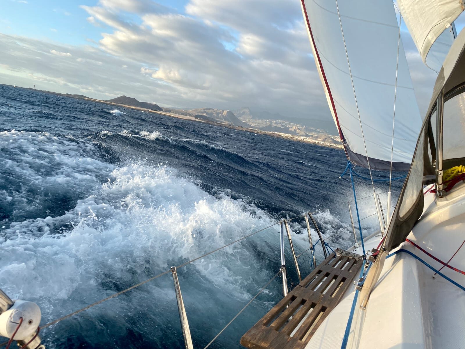 Segelboot durchpflügt raue Wellen vor einer Küste mit Vulkanbergen, Schaumkronen spritzen über den Bug.